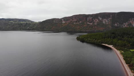 Aerial-View-Of-Beach-Shoreline-At-Loch-Ness-With-Mountain-Landscape-In-Background