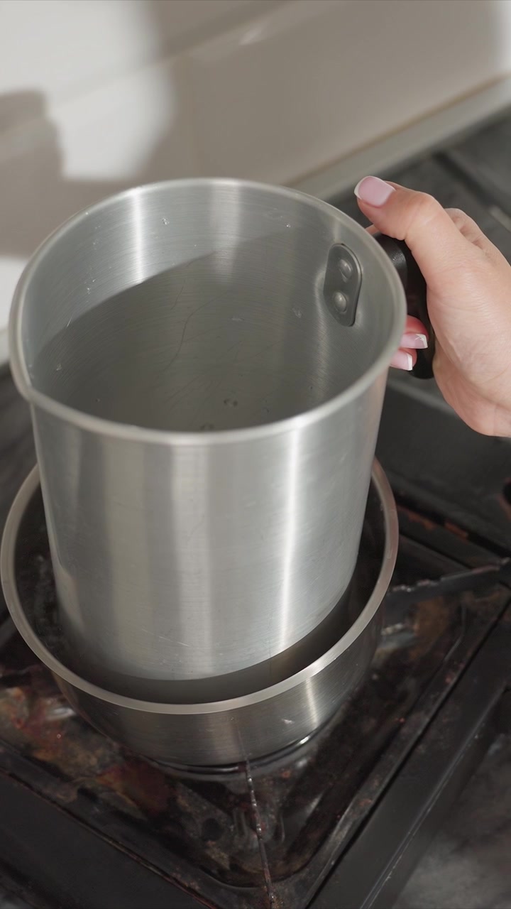 Primo piano di acqua che bolle in una pentola di acciaio inossidabile su un fornello elettrico. Una mano posiziona e sistema un altro contenitore metallico vuoto sopra il liquido che bolle, indicando una manipolazione attenta durante il riscaldamento.