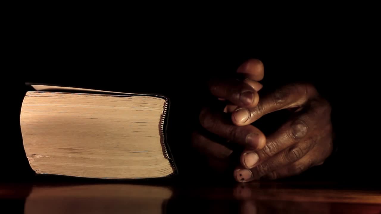 Vidéo de stock: Homme caribéen priant, les mains jointes, avec une Bible, sur fond noir.