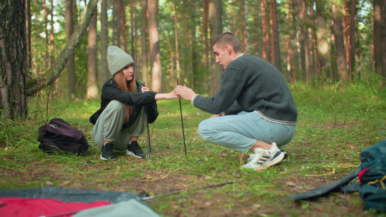 Young couples squatting preparing tent in forest as lady hands pole to partner with care, surrounded by trees and scattered gear, expressing warmth, teamwork, and love