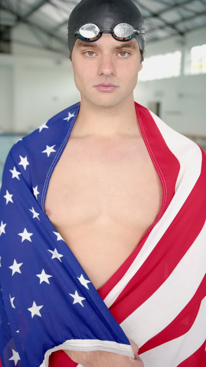 Vertical video: Swimmer wearing goggles and American flag, standing confidently in indoor pool