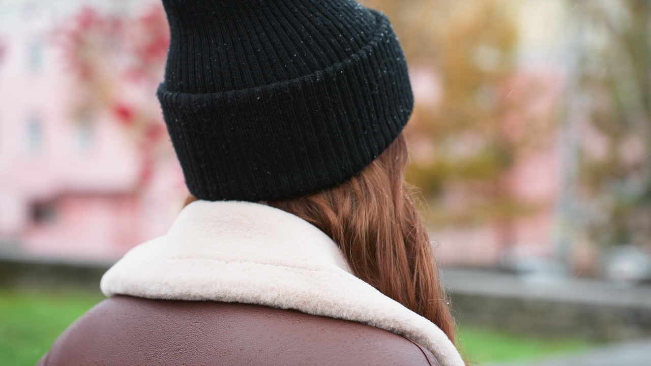 Close back view of young girl wearing black knit cap and brown shearling jacket walking outdoors during overcast day, blurred urban park background adds calm peaceful atmosphere