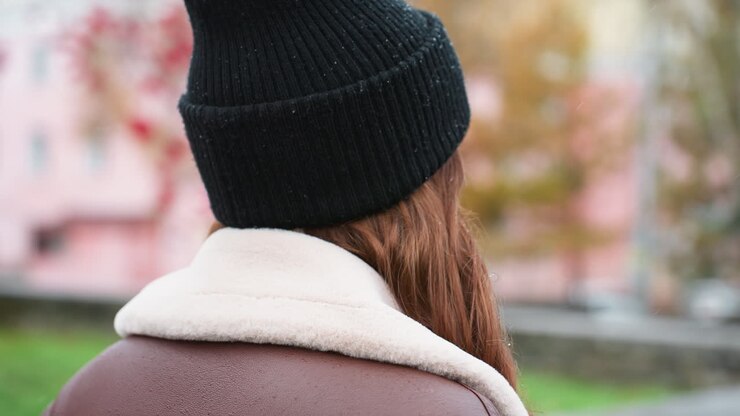 Primo piano da dietro di una giovane ragazza che indossa un berretto di maglia nero e una giacca marrone in shearling mentre cammina all'aperto in una giornata nuvolosa; lo sfondo sfocato del parco urbano aggiunge un'atmosfera calma e tranquilla.