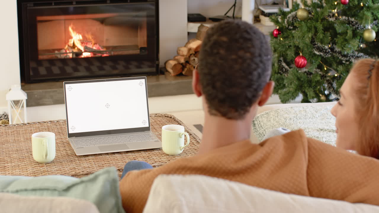 Diverse couple relaxing by fireplace with laptop and Christmas tree nearby, copy space