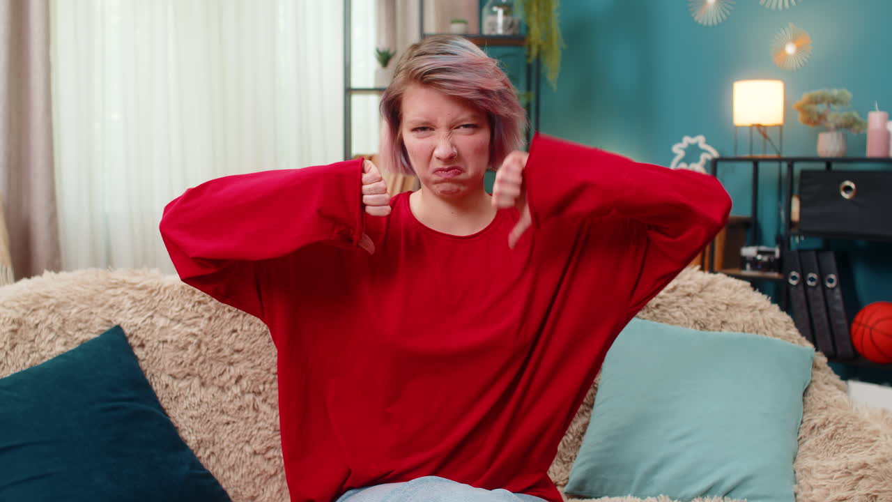 Young woman showing thumbs down gesture negative disapproval disagreement rejection reaction at home