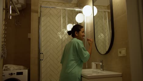 vista lateral de feliz morena chica india en pijama verde se cepilla los dientes mientras se prepara para la cama en el baño en un apartamento moderno por la noche