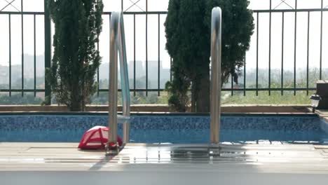 Close-up-of-a-red-baseball-hat-next-to-the-swimming-pool-ladder,-blurred-city-panorama-visible-in-background