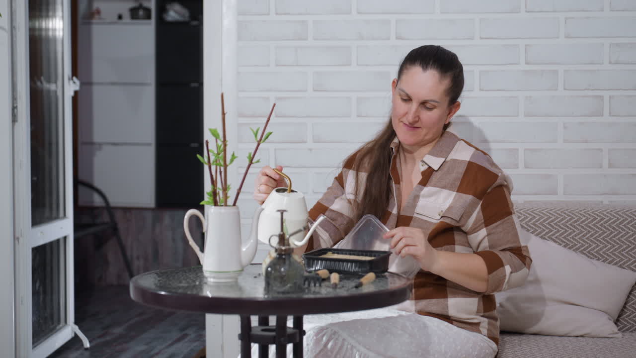 Cheerful lady seated by round glass table opening disposable food container and pouring water from jug into tray among potted plants in cozy indoor setting with white brick wall and warm lighting
