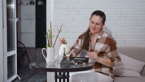 cheerful lady seated by round glass table opening disposable food container and pouring water from jug into tray among potted plants in cozy indoor setting with white brick wall and warm lighting