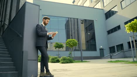 stylish bearded businessman in formal business suit standing working with tablet in hands on background modern office building outside.