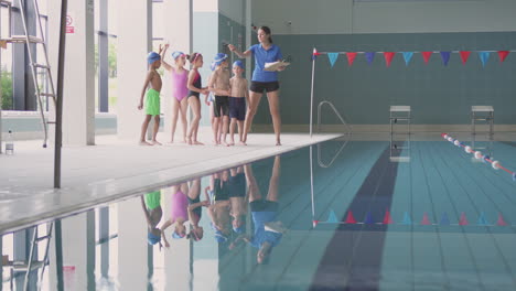 Female-Coach-Walks-Children-In-Swimming-Class-Along-Edge-Of-Indoor-Pool