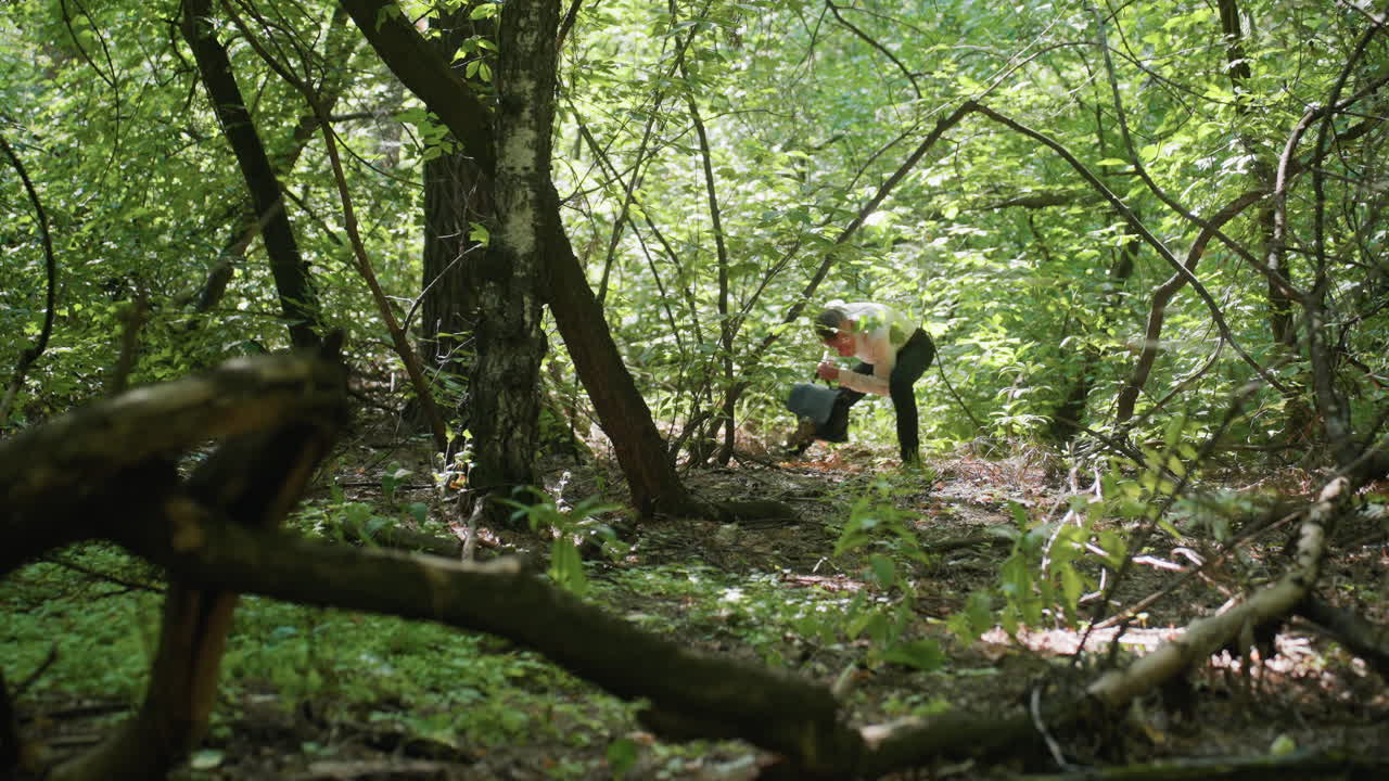 Man carrying backpack bending while walking carefully through dense green forest under daylight, moving between branches and trees surrounded by natural greenery expressing exploration