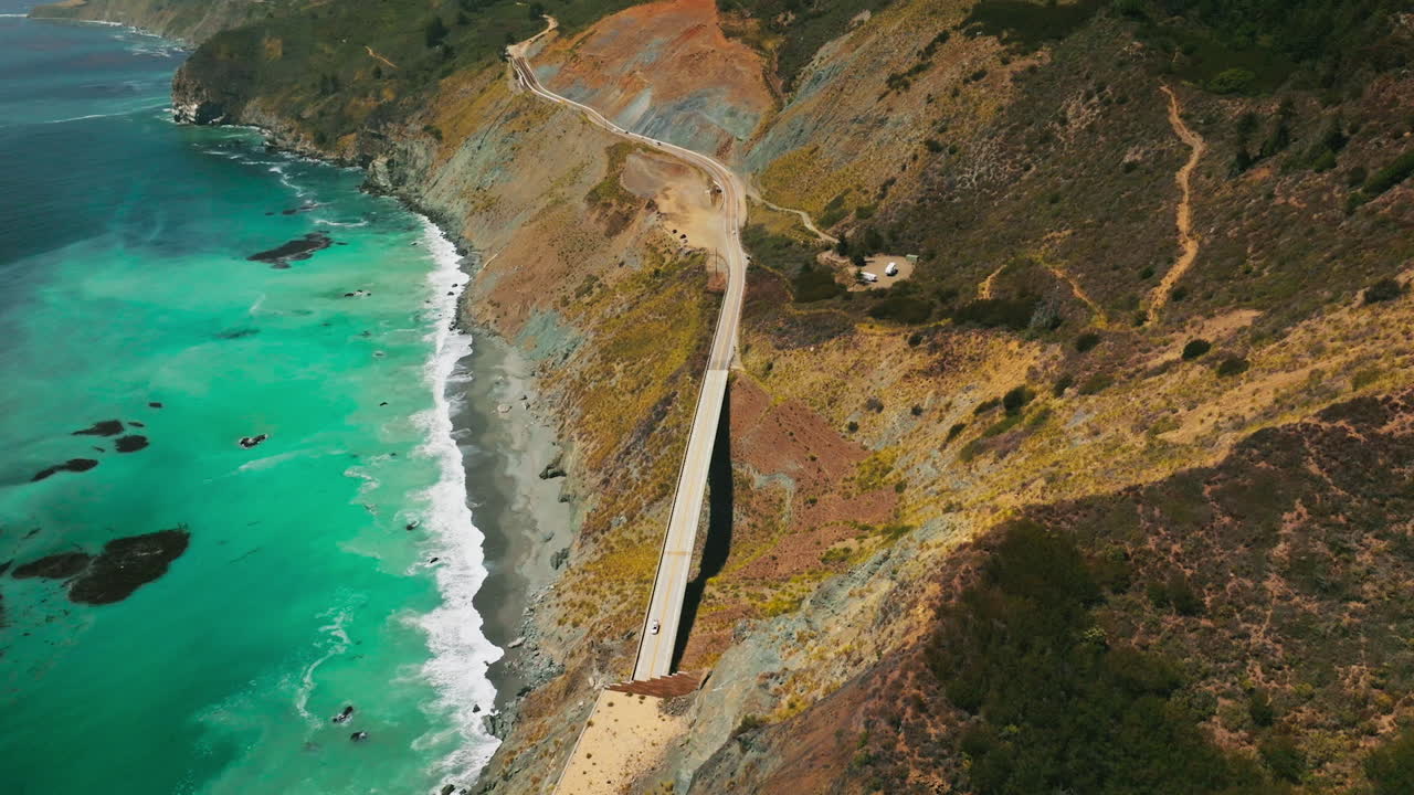 Flight over the highway in the sloping mountains of California coast. Beautiful azure ocean splashing by the rocks. Aerial view.