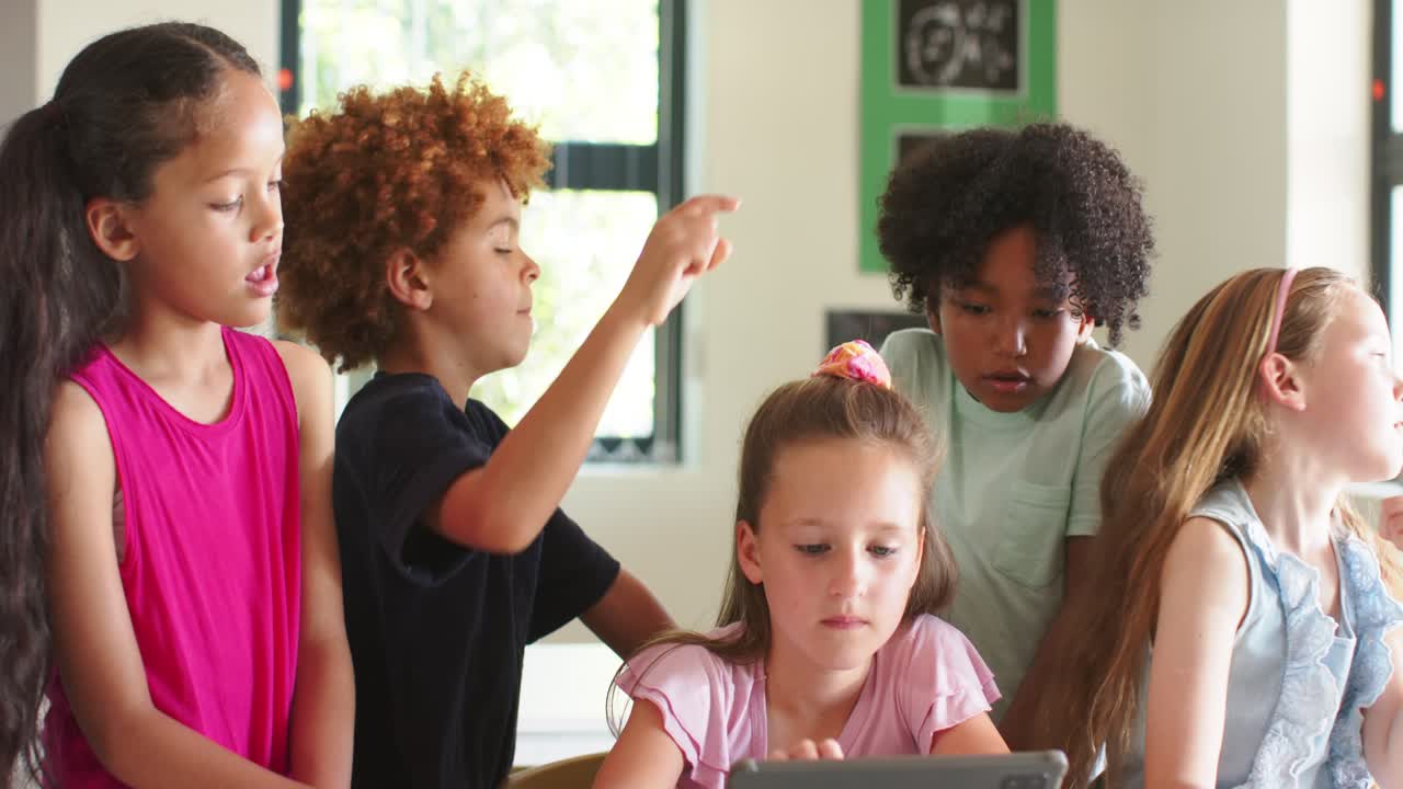 Five schoolkids leaning over tablet at table, girl tapping screen while boy pointing upward