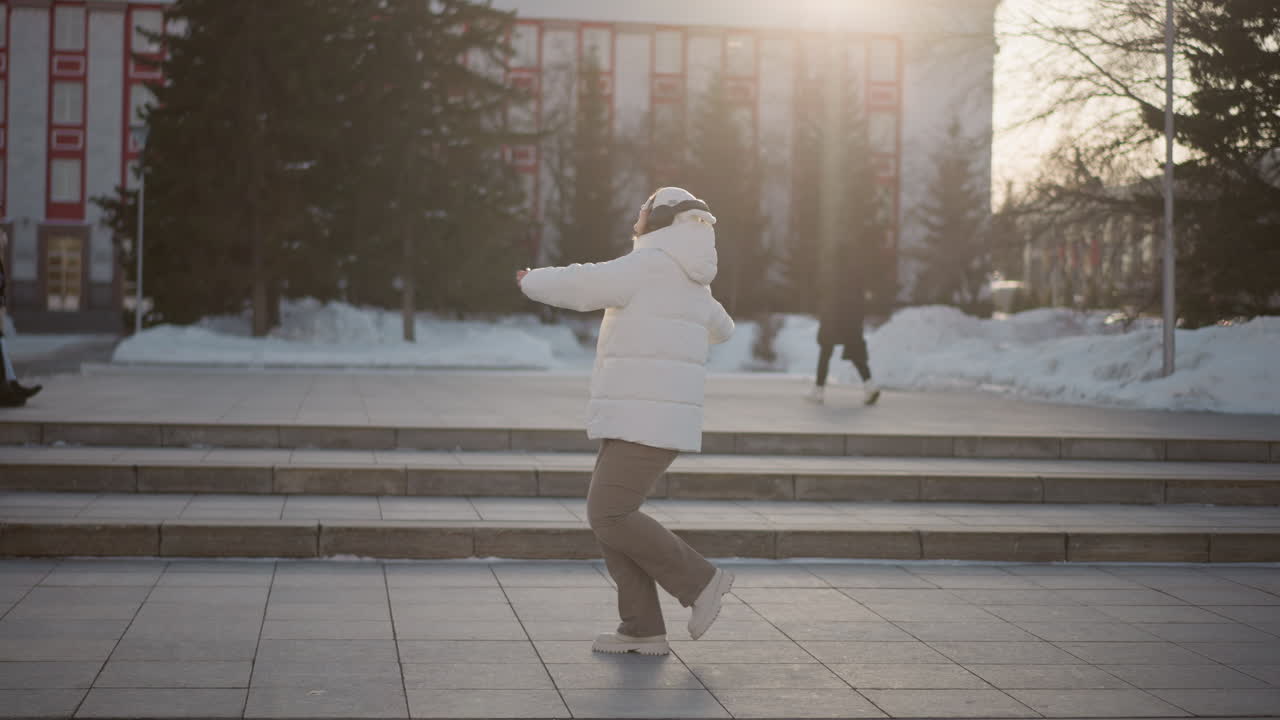 Young woman in winter coat and headphones spins gracefully on snowy step pavement during golden hour, immersed in dance while people walk in background near trees under soft sunlight