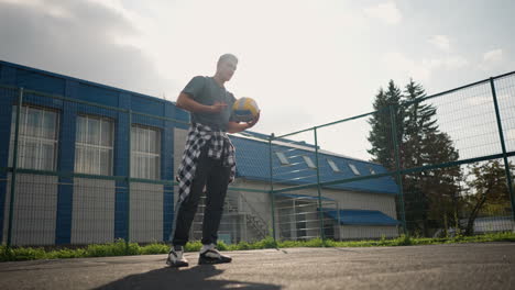athlete during training session bouncing volleyball, rotating it in hand before slamming it, background shows greenery and building, captured outdoors in a sport arena environment