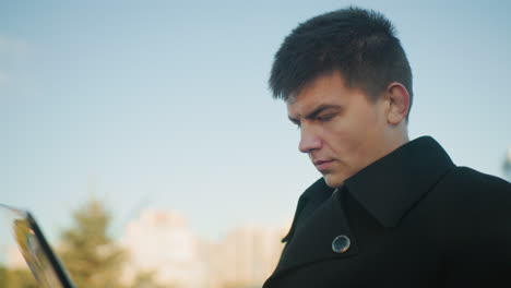 man in black coat focused on operating laptop outdoors in urban environment, blur background featuring modern office building and city elements in background