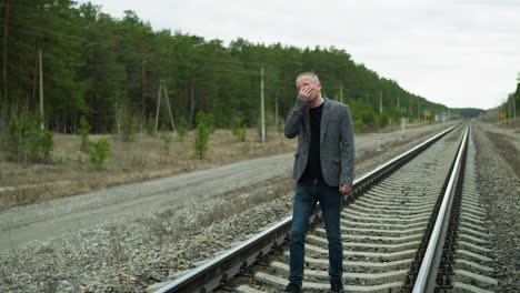 a close-up view of a middle-aged man in a grey suit and black shirt walking along railway tracks in a forested area, the man covers his face with one hand, appearing deep in thought