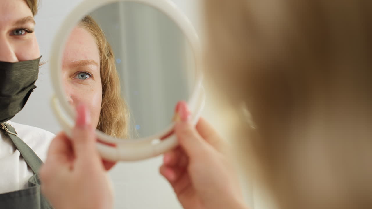 Client examines freshly shaped eyebrow in mirror with visible reflection confirming satisfaction. Beautician wearing black face mask stands beside as client evaluates result in softly lit salon environment
