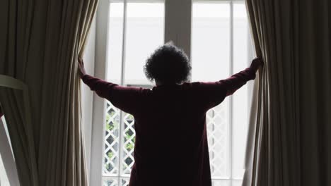 vista trasera de una mujer afroamericana mayor abriendo las cortinas de la ventana en casa