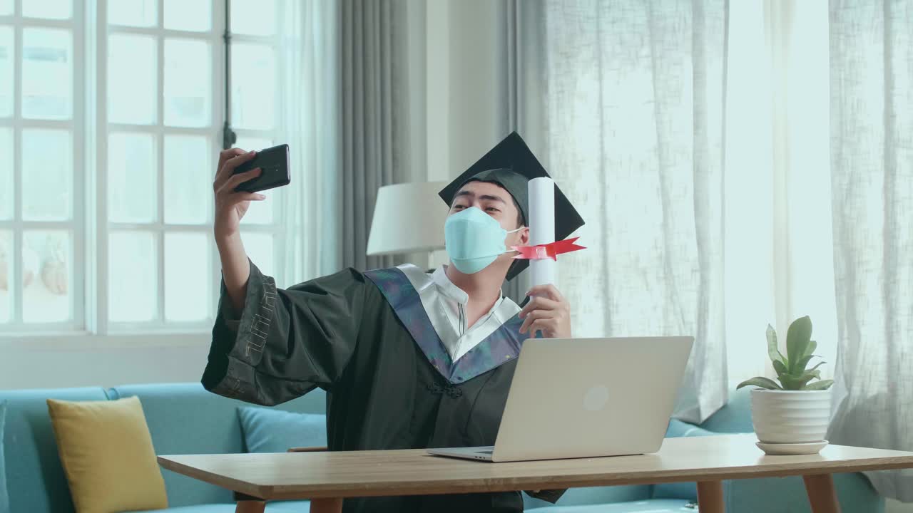 Giovane uomo asiatico che indossa una mascherina protettiva, si scatta un selfie mentre indossa una toga e un tocco da laureato, con in mano un certificato universitario in salotto.