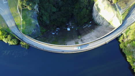 Luftaufnahme-Eines-Autos,-Das-über-Einen-Damm-Mit-Blauem-Wasser-Und-Grünem-Wald-Fährt