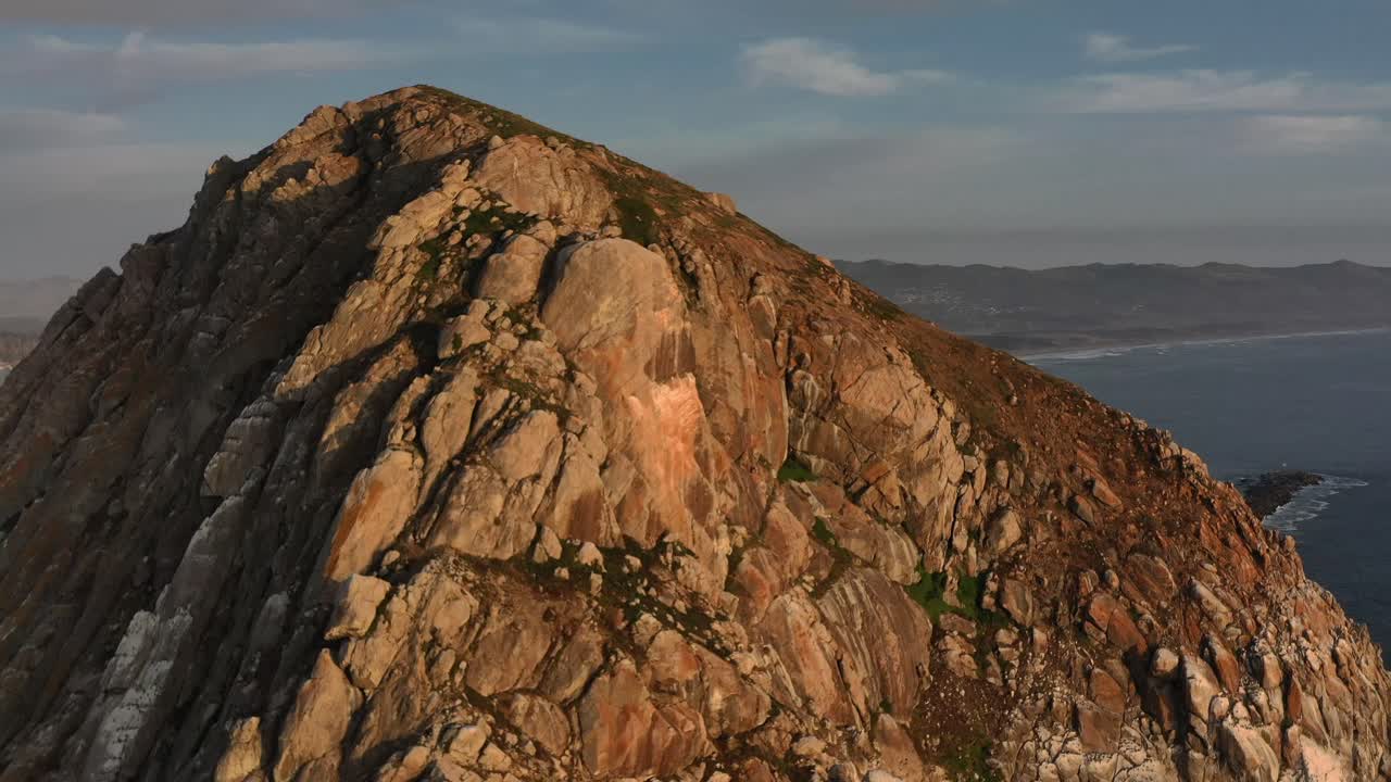 Panoramica aerea con drone sopra Morro Bay Rock al tramonto, rivelando la baia retrostante e una barca da pesca ancorata nel porto.