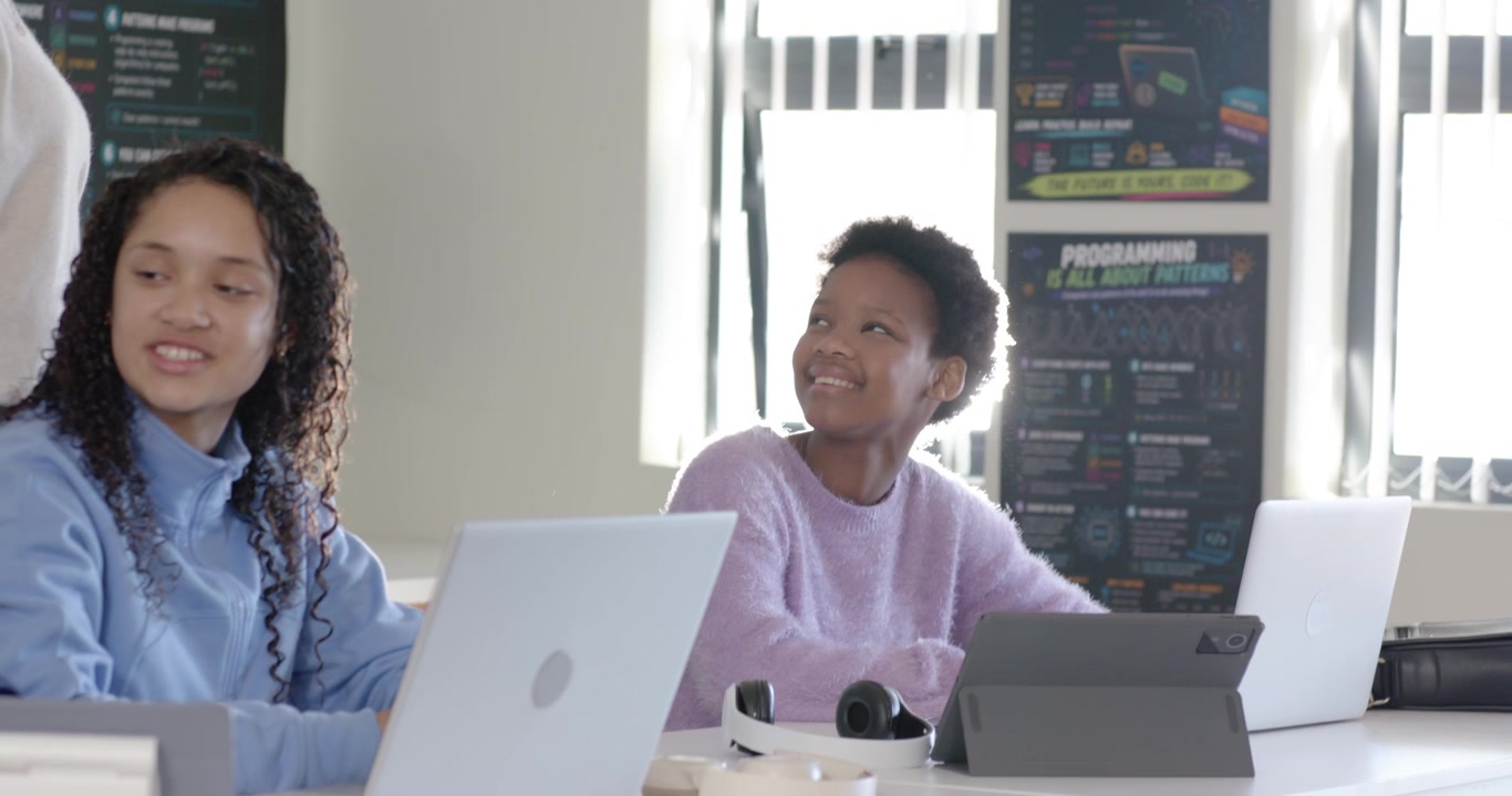 African American female teens using laptops looking up in lab while Indian teacher walking to help
