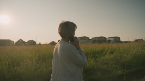 side view of woman talking on phone while walking close to grassy field during golden hour, distance buildings visible in background, sunlight casting warm glow over rural landscape