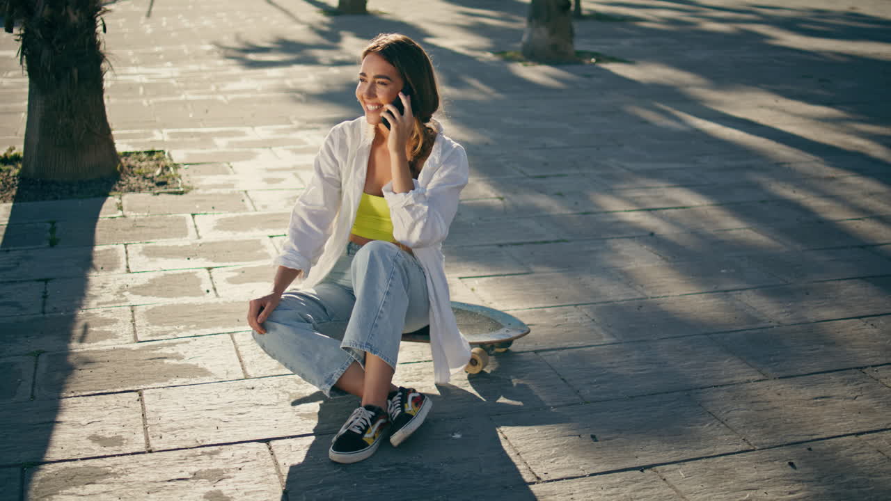 estudiante sonriente hablando por teléfono móvil en el callejón. mujer feliz descansando en longboard