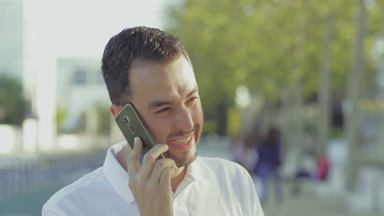 joven sonriente emocional hablando en el teléfono inteligente al aire libre.