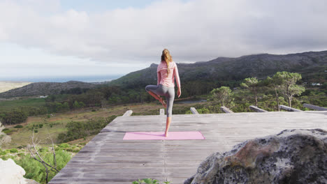 vista trasera de una mujer caucásica practicando yoga de pie en una pierna estirándose en la cubierta en la ladera de la montaña