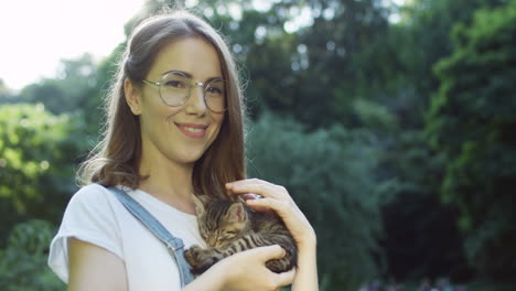 close-up view of a caucasian woman in glasses holding and petting kitty cat while smiling at camera in the park on a summer day