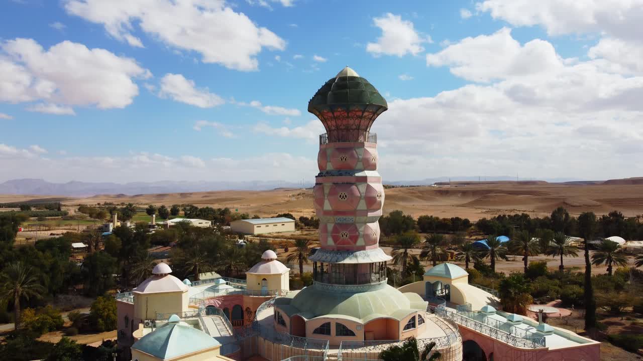 Drone circles around Neot Smadar Arts Center cooling tower under clear blue sky with organic agricultural landscape, Arava desert in background