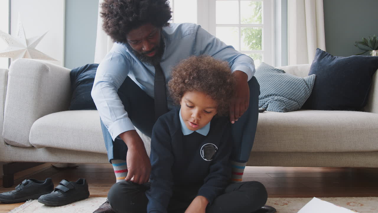 Front view of pre-teen boy in school uniform sitting on the floor at home doing his homework helped by his father, sitting behind him on the sofa, zoom out shot