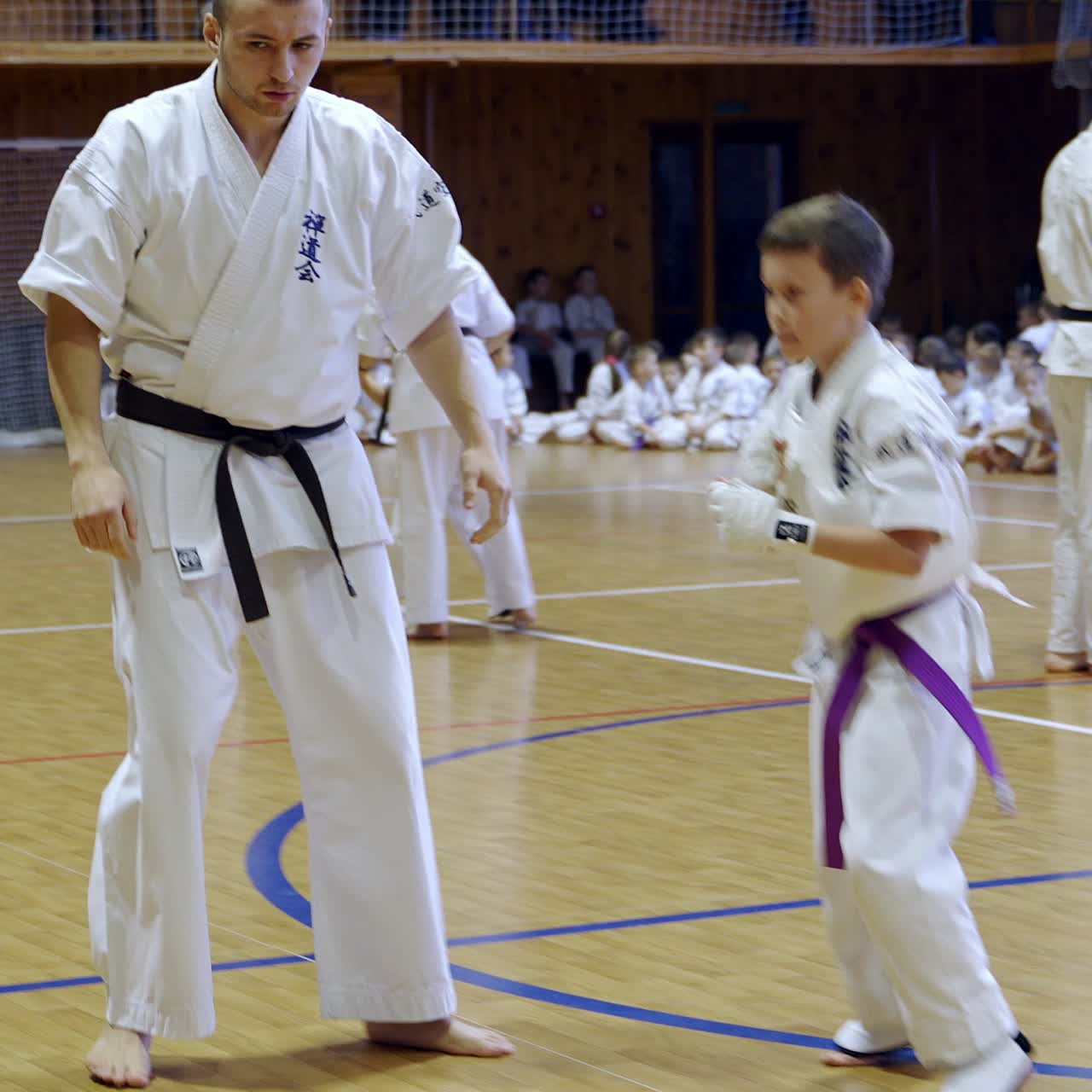 Hai cậu thiếu niên đang tập luyện karate dưới sự hướng dẫn của huấn luyện viên nam. Các cặp vận động viên khác giao đấu ở phía sau.
