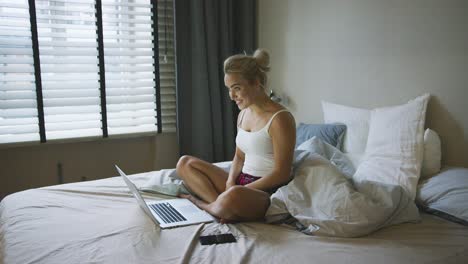 mujer complacida sentada en la cama cerca de la computadora portátil