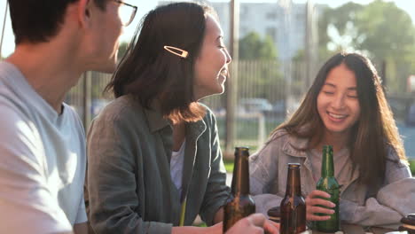 group of three japanese friends talking and drinking beer while sitting at table outdoors in a sunny day