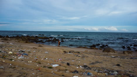 Drone-approach-female-tanned-surfer-standing-on-tropical-beach-with-plastic-waste-garbage-trash-toxic-pollution,-climate-change-concept