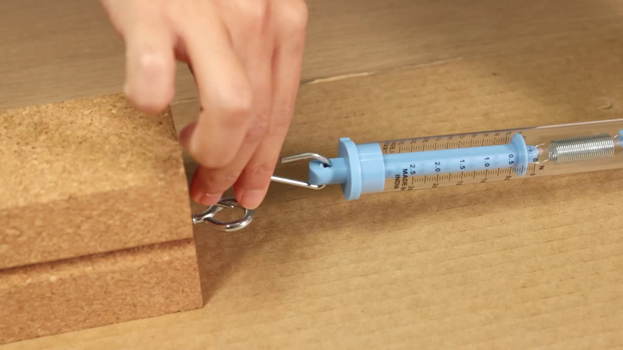 A hand uses a spring scale to measure friction on a wooden block. Close-up, well-lit, detailed view of the process