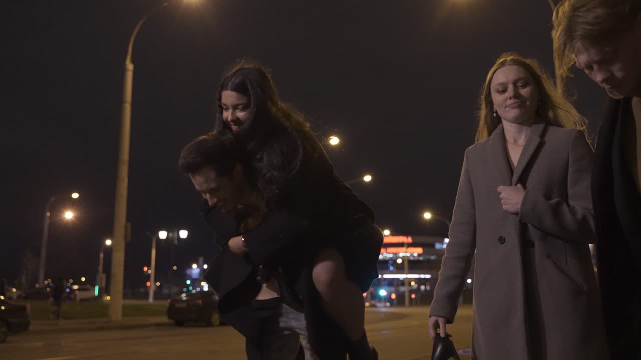 Two couples of friends dressed in elegant clothes walking in the street street while talking and laughing after New Year's party, one of the girls is perched on her boyfriend's back