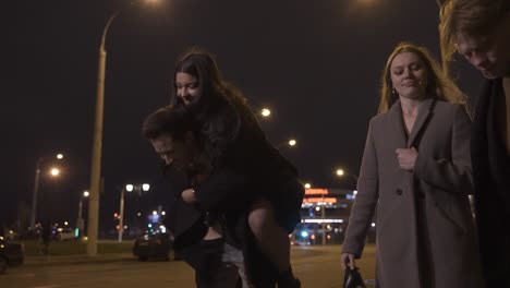 Two couples of friends dressed in elegant clothes walking in the street street while talking and laughing after New Year&#039;s party, one of the girls is perched on her boyfriend&#039;s back