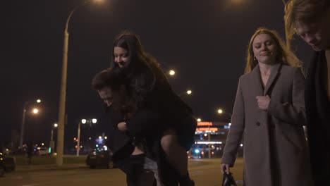 two couples of friends dressed in elegant clothes walking in the street street while talking and laughing after new year's party, one of the girls is perched on her boyfriend's back