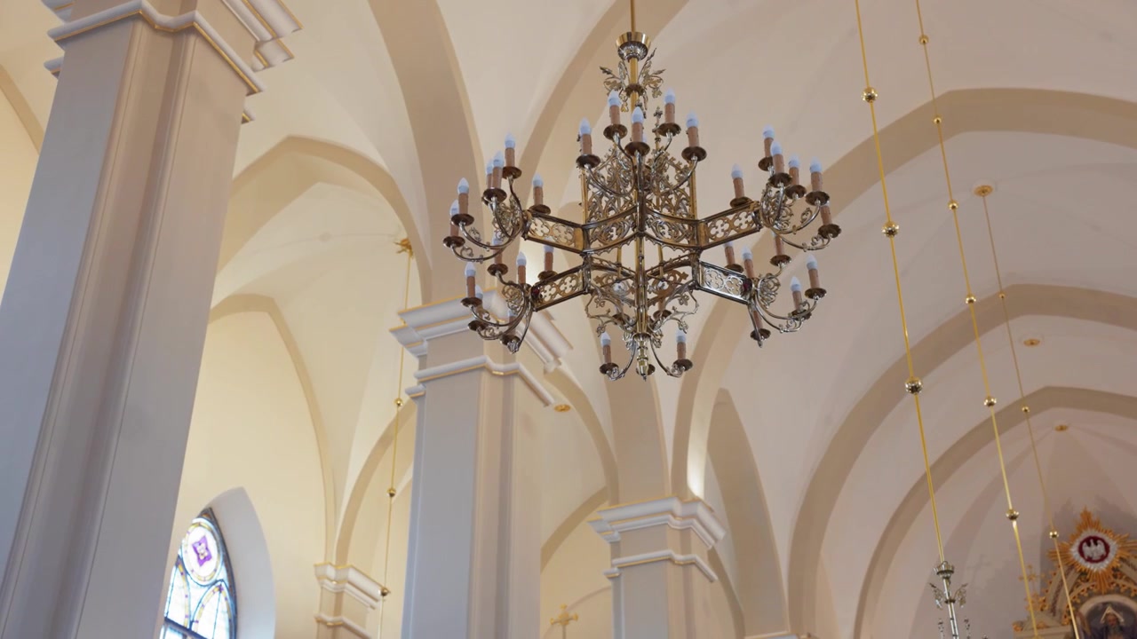 Vista dettagliata di un lampadario ornato appeso a soffitti alti ad arco color crema all'interno di una chiesa storica. La luce soffusa del giorno riempie lo spazio, evidenziando la bellezza architettonica in un'inquadratura ampia e stabile.