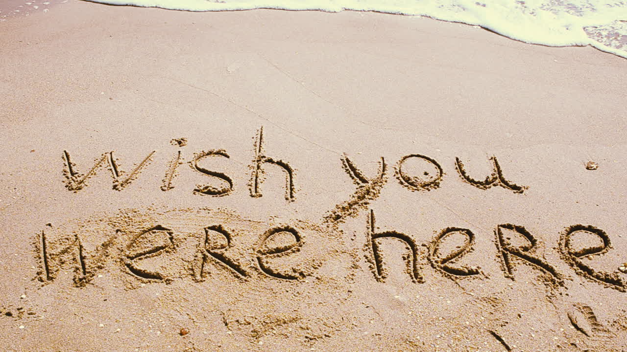 Testo concettuale 'wish you were here' scritto a mano sulla sabbia di una spiaggia.