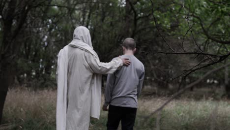 Robed-man-stands-in-forest-looking-at-a-young-teen-boy