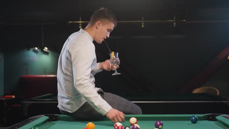 gentleman in white shirt and checkered trousers sits on green pool table, holding glass of lemon drink while rolling billiard ball. stylish setting with ambient lighting and modern pool hall