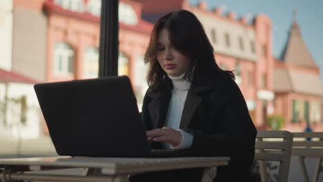 remote worker seated outdoors at mall working on laptop with blurred background featuring urban buildings and someone moving in the distance