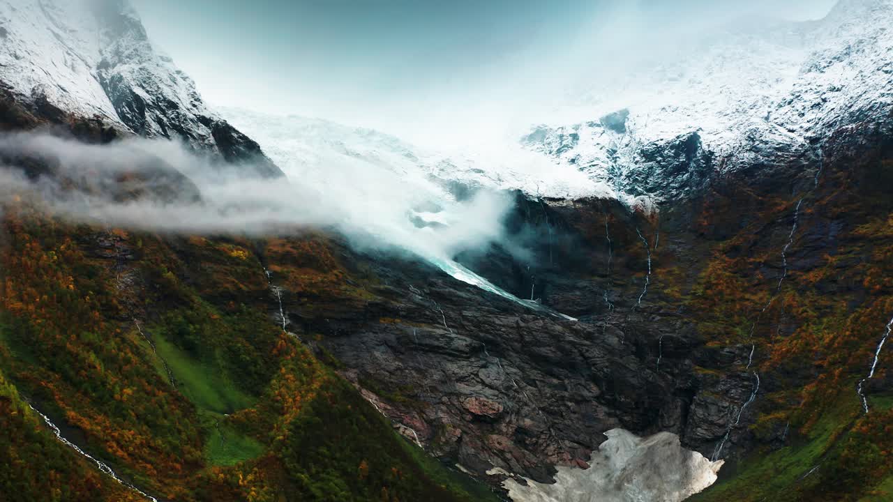 Una montagna coperta da ghiacciai è avvolta nella nebbia, con cascate che scendono a picco da ripide scogliere e il fogliame autunnale.