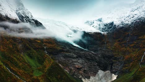 a glacier-covered mountain is shrouded in mist, with waterfalls cascading down steep cliffs and autumn foliage